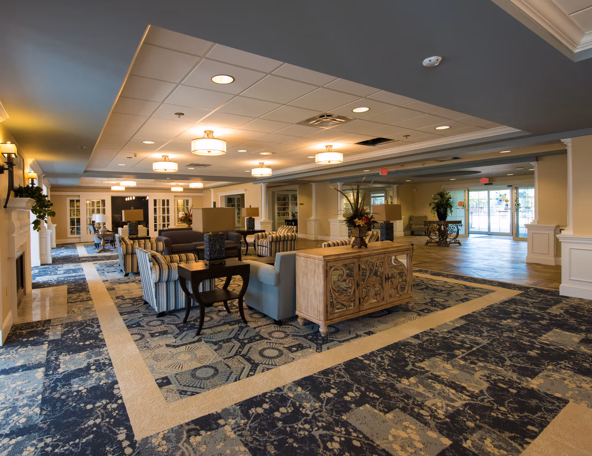 Spacious and well-lit senior living facility common area with patterned carpet, multiple seating arrangements including striped and solid armchairs, wooden tables with lamps, decorative plants, and large windows near the entrance allowing natural light.