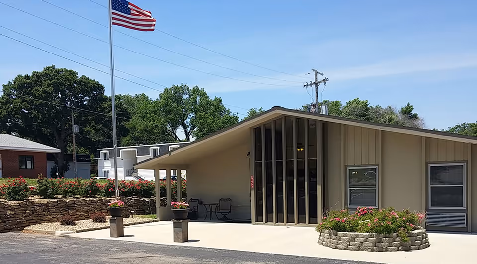 Exterior view of a single-story building with a slanted roof, large vertical windows, and a covered entrance area. There are flower beds with pink flowers and an American flag on a tall flagpole in front of the building. Trees and other buildings are visible in the background under a clear blue sky.