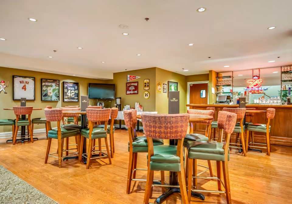 Interior view of a lounge area with wooden floors, several round tables surrounded by high-back chairs with green cushions, a bar counter with stools, a TV mounted on the wall, and sports memorabilia including framed jerseys and pennants displayed on the walls.