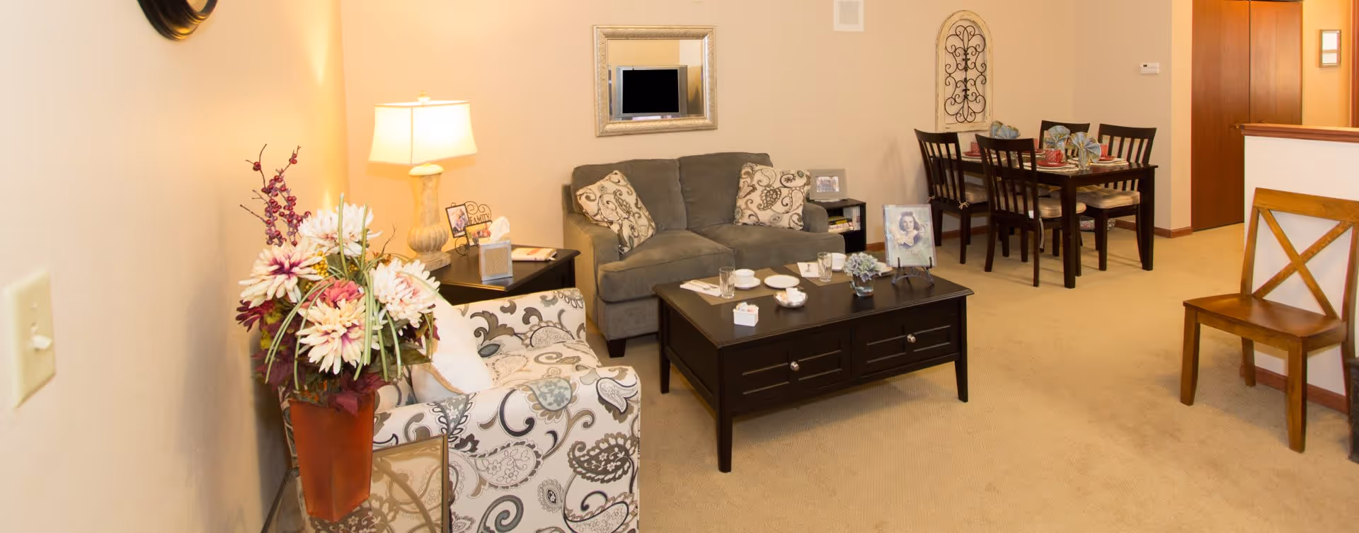 A cozy living room and dining area in a senior living facility. The living room features a patterned armchair, a gray loveseat with decorative pillows, a dark wooden coffee table with cups and a small plant, and a side table with a lamp and framed photos. The dining area has a dark wooden table set with plates, napkins, and cups, surrounded by four chairs. The walls are beige, and there is a mirror and decorative wall art. The carpet is light beige.