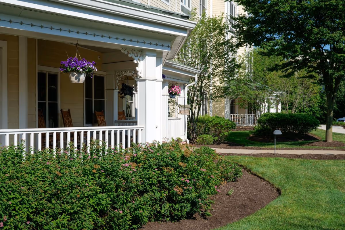 View of the exterior of a senior living facility showing a porch with rocking chairs and hanging flower baskets. The building is light-colored with white trim and surrounded by well-maintained landscaping including bushes, trees, and a green lawn under a clear blue sky.