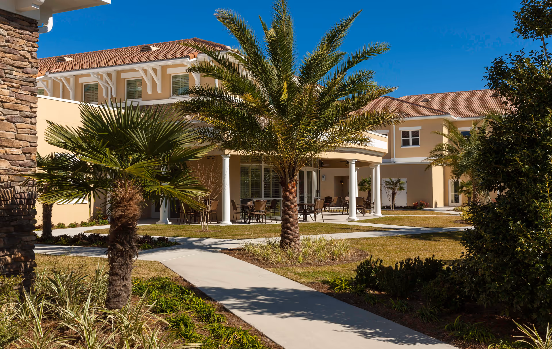 Outdoor view of HarborChase of Villages Crossing showing a beige two-story building with a tiled roof, surrounded by palm trees and landscaped greenery. There is a paved walkway leading to a covered patio area with tables and chairs under a clear blue sky.