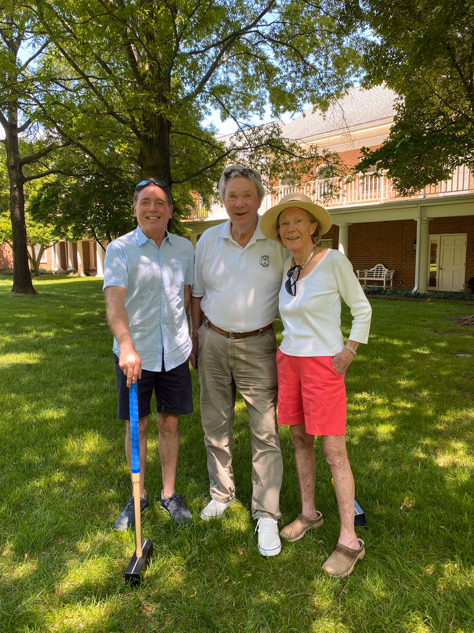 Three people standing on a grassy lawn under trees in front of a brick building with a covered porch. One man on the left is holding a croquet mallet, the man in the middle is wearing a white polo shirt and khaki pants, and the woman on the right is wearing a white long-sleeve shirt, red shorts, a sun hat, and brown slip-on shoes.