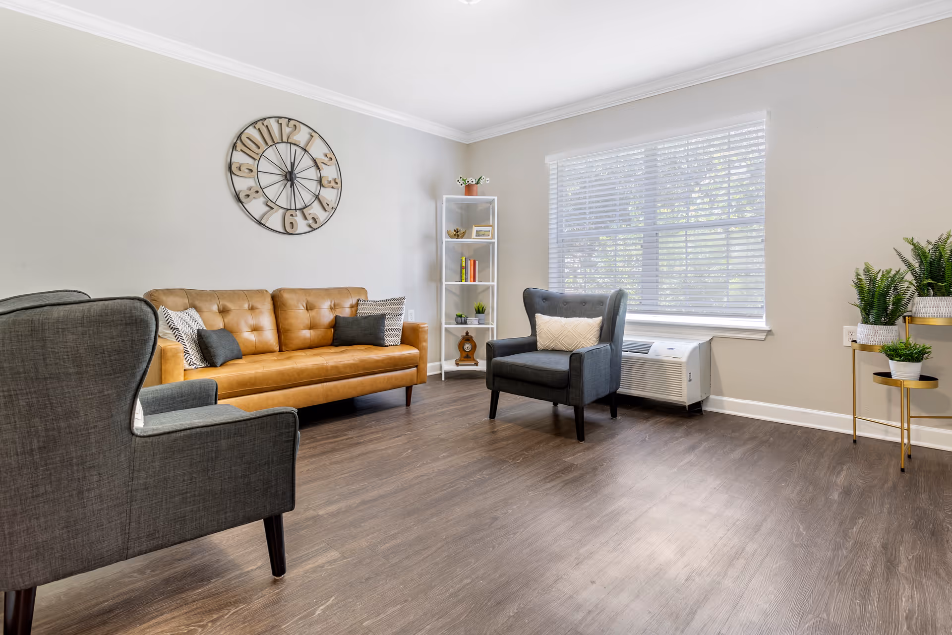 A bright and spacious living room with a tan leather sofa adorned with pillows, two gray armchairs, a large wall clock, a white shelving unit with decorative items, a window with blinds, and several potted plants on a small gold and white stand.