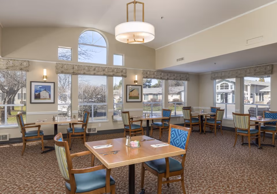A bright dining room with several wooden tables and chairs arranged neatly. The chairs have blue and green patterned cushions. Large windows with blinds and valances allow natural light to fill the room, showing an outdoor view of trees and buildings. The walls are light-colored with framed artwork and wall sconces, and a modern ceiling light fixture hangs above.
