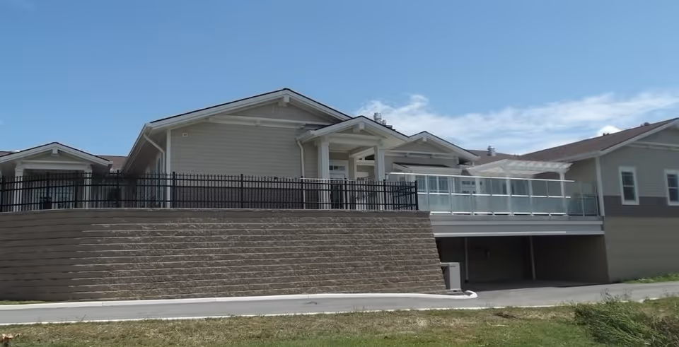 Exterior view of a single-story building with beige siding, a sloped roof, and a raised patio area enclosed by a black metal fence and glass railing under a blue sky with some clouds.