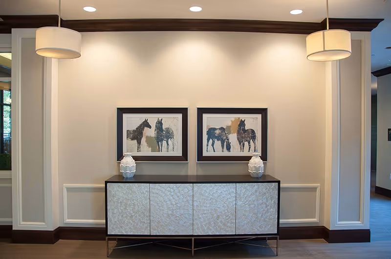 A hallway or lobby area with a decorative sideboard featuring a textured front and two white vases on top. Above the sideboard are two framed artworks depicting horses. The walls are light-colored with dark wood trim, and two pendant lights hang from the ceiling on either side of the sideboard.