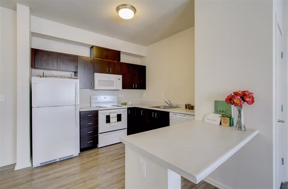 A modern kitchen with white appliances including a refrigerator, stove, microwave, and dishwasher. The kitchen has dark wood cabinets, a light countertop with a small overhang for seating, and a vase with red flowers on the counter. There is also a green folder labeled 'LARC' and a small welcome card on the counter.