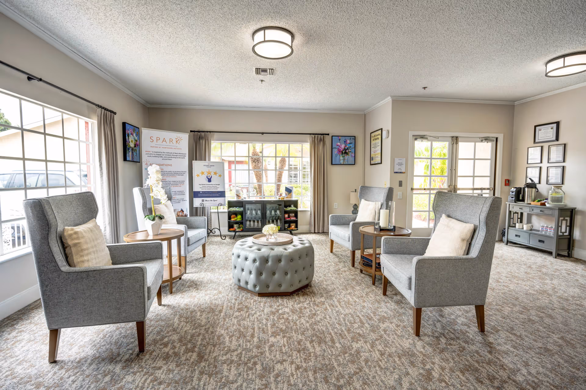 Bright memory-care lounge with four gray armchairs arranged around a tufted ottoman, side tables, large windows, and decorative accents.