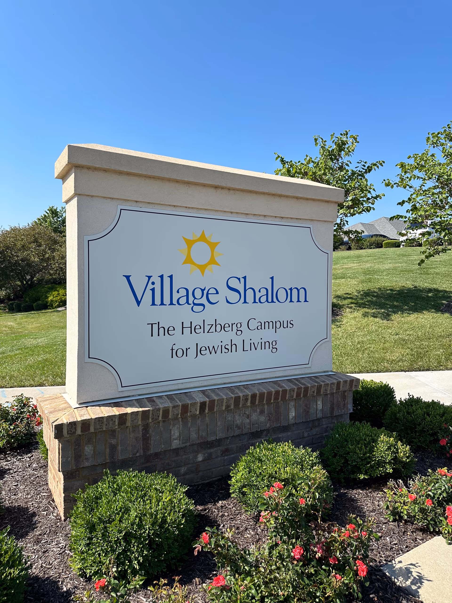 Outdoor stone sign for Village Shalom, The Helzberg Campus for Jewish Living, surrounded by green bushes and red flowers with a clear blue sky in the background.