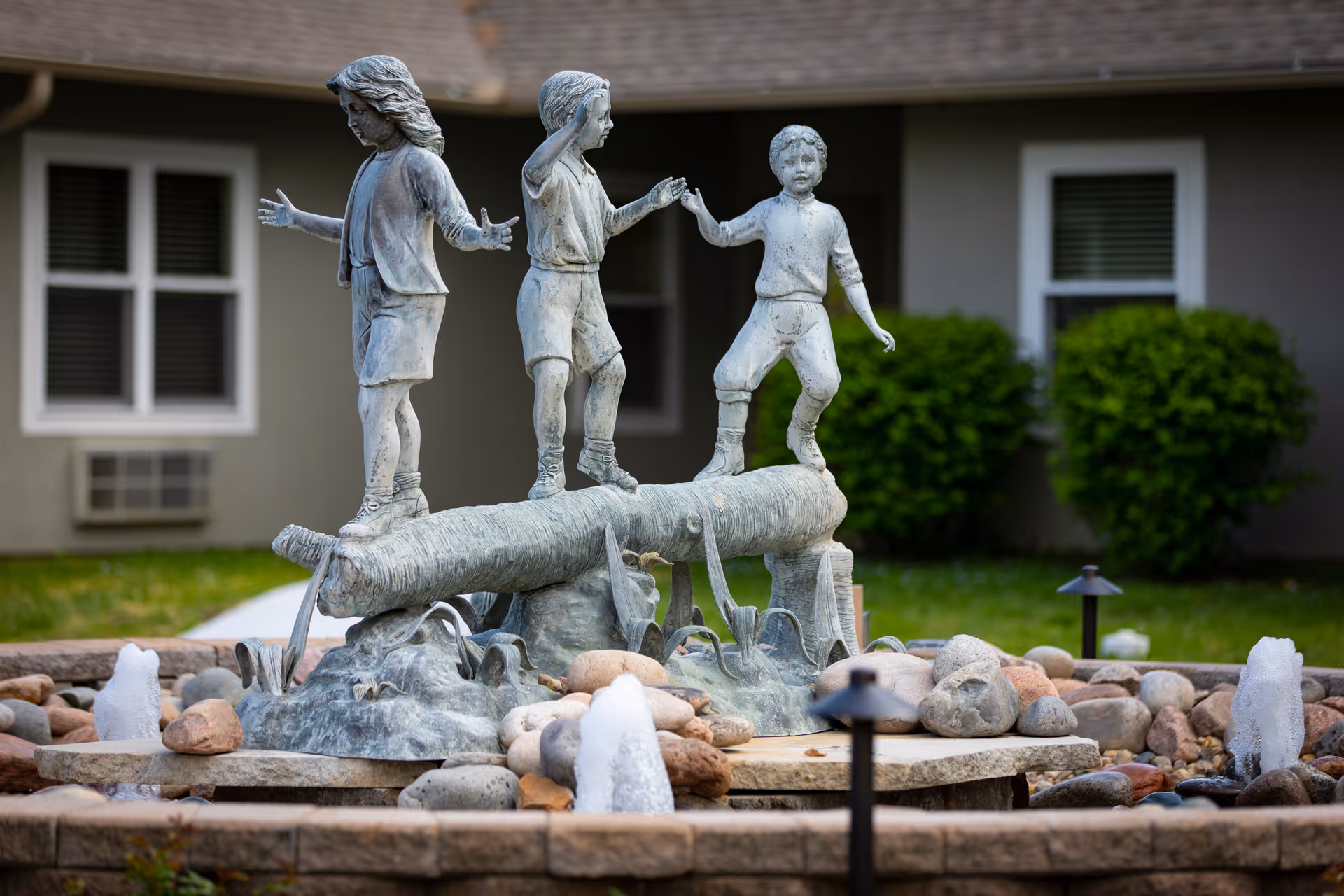 A bronze statue of three children balancing on a log, surrounded by a circular stone water fountain with small water jets and rocks, set in an outdoor garden area with a building and green bushes in the background.