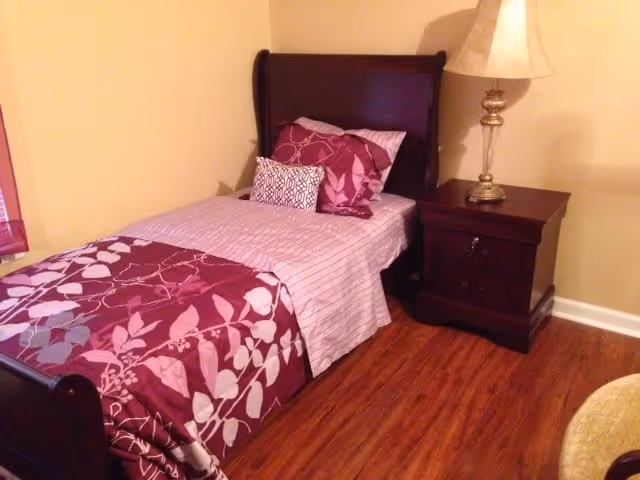 A single bed with a dark wooden frame, made up with a purple and white floral patterned comforter, striped sheets, and multiple pillows. Next to the bed is a dark wooden nightstand with a decorative lamp on top. The room has wooden flooring and beige walls.