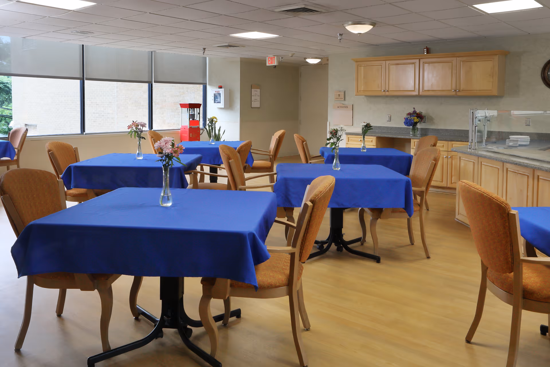 Dining room with tables covered in blue tablecloths, chairs, small vases of flowers, and a serving counter in a senior living facility.