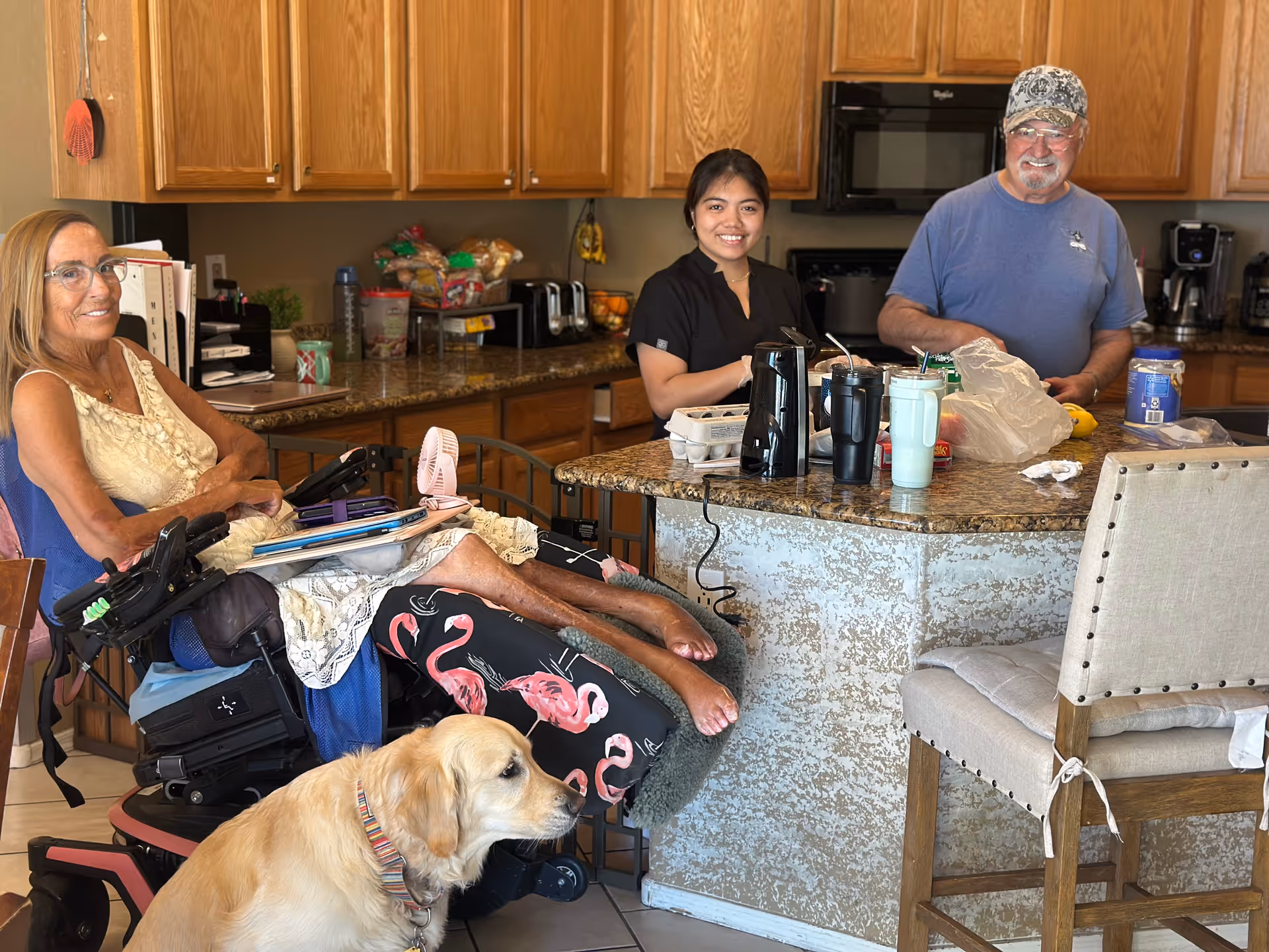 A woman in a wheelchair with flamingo-patterned pants and a golden retriever dog sits at a kitchen island with a smiling young woman and an older man standing behind the counter in a kitchen with wooden cabinets and granite countertops.