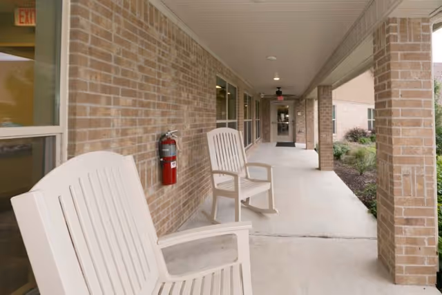 Covered outdoor walkway with beige brick walls and white plastic chairs along the side. There is a fire extinguisher mounted on the wall and a door at the end of the walkway. Some landscaping with plants is visible on the right side.