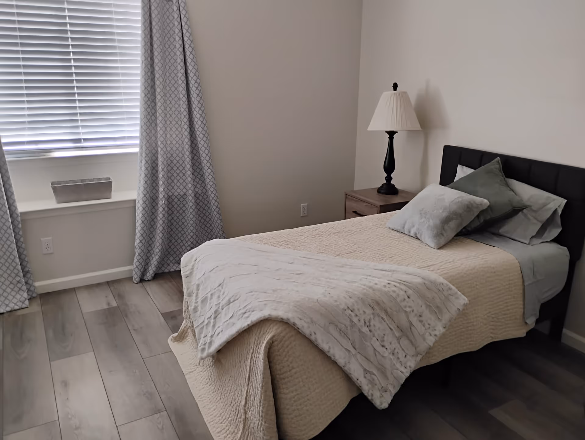 A neatly made single bed with beige bedding and two pillows, one green and one light gray, in a simple bedroom. Next to the bed is a wooden nightstand with a black lamp with a white lampshade. A window with closed blinds and patterned gray curtains is on the left side of the room. The floor is light-colored wood.