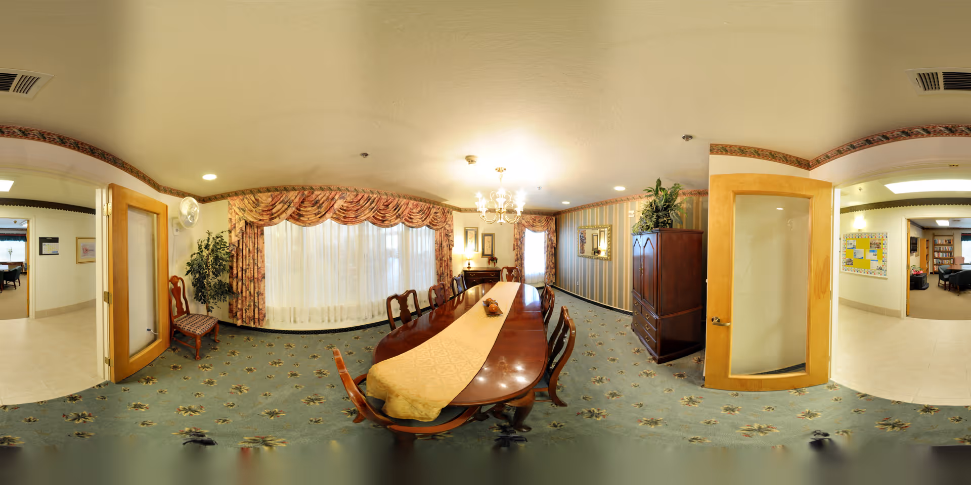A formal dining room with a long wooden table covered by a beige table runner and surrounded by wooden chairs. The room has floral carpet, patterned curtains over large windows, a chandelier hanging from the ceiling, and a wooden cabinet with a plant on top. Two open wooden doors lead to adjacent rooms with light-colored flooring and walls.
