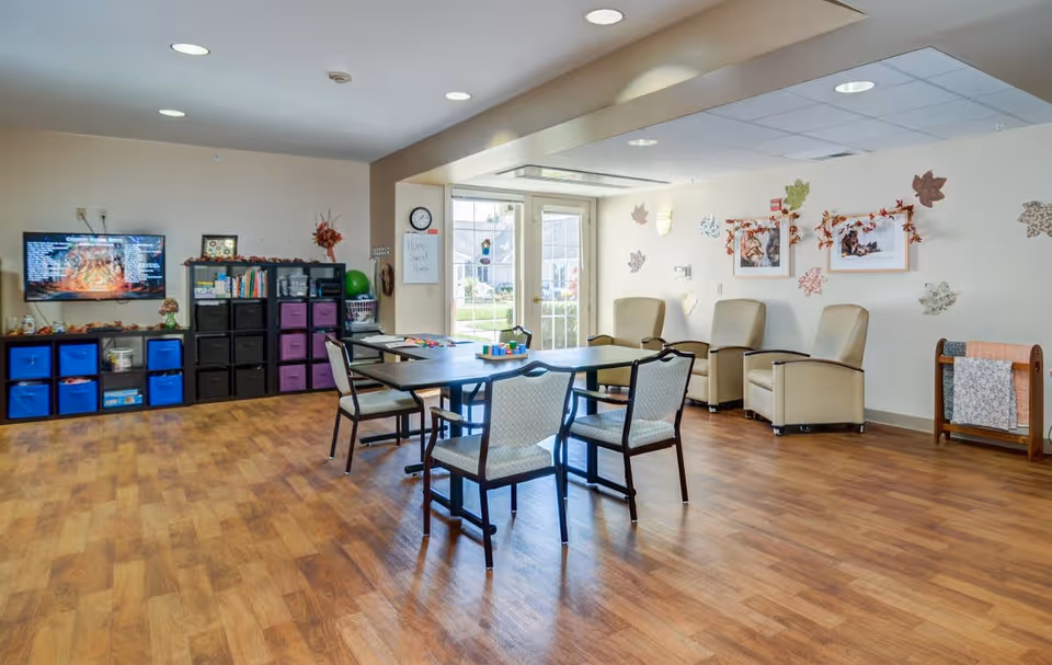 A bright senior living community room with wooden flooring, a table with four chairs in the center, three beige armchairs against the wall, and a shelving unit with colorful storage bins and books. The walls are decorated with autumn-themed leaves and framed pictures. A TV is mounted on the wall to the left, and a door with windows leads outside.