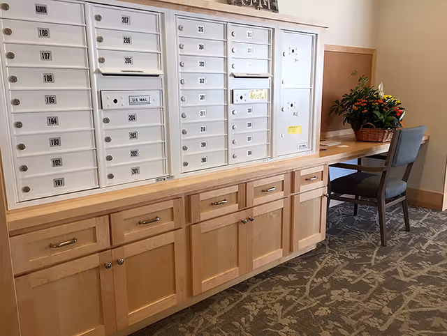 Interior view of a mailroom area in a senior living facility featuring multiple white mailboxes mounted above wooden cabinets. To the right, there is a wooden desk with a chair and a basket of flowers placed on top. The floor is carpeted with a patterned design.