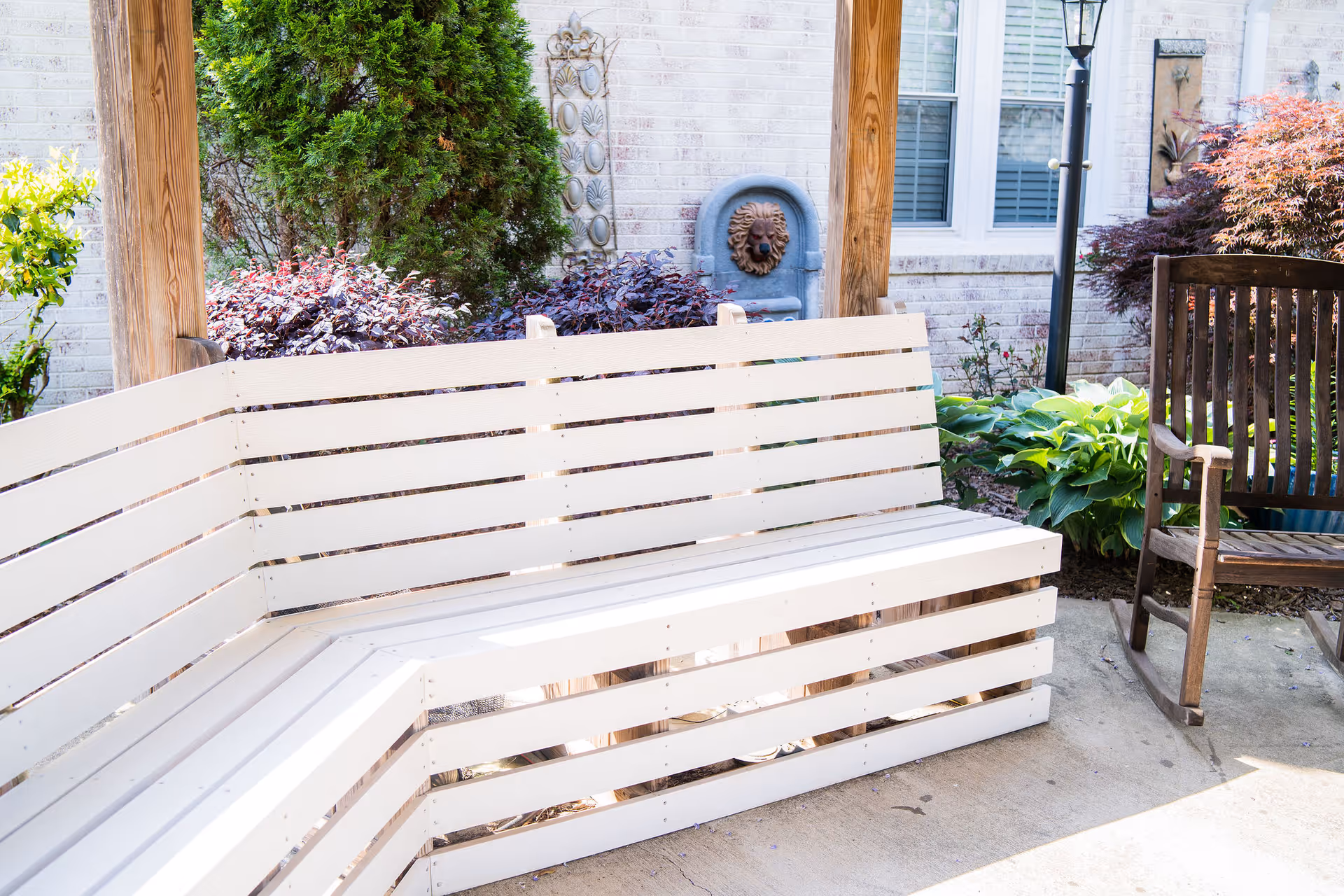 Outdoor seating area with a white wooden bench and a wooden rocking chair on a concrete patio. The background features a brick wall with windows, a decorative lion head water fountain, various green plants, and a lamppost.