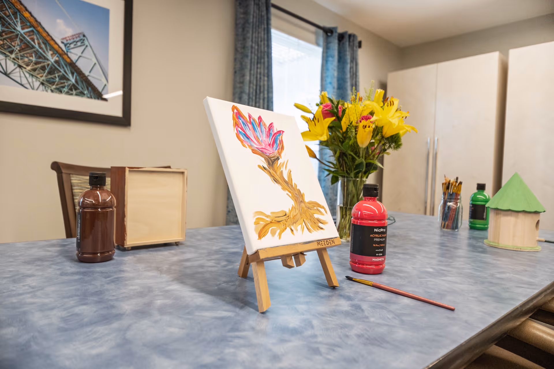 A table with painting supplies including a small canvas on an easel featuring a colorful flower painting, bottles of paint, paintbrushes, a wooden box, and a vase of yellow flowers in a room with blue curtains and a framed picture on the wall.