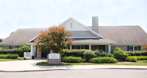 Exterior view of a single-story building with a gabled roof, surrounded by greenery and bushes. A tree with red and green leaves stands in front of the building near a sign that reads 'Sunrise House'. The sky is clear and the area appears well-maintained.