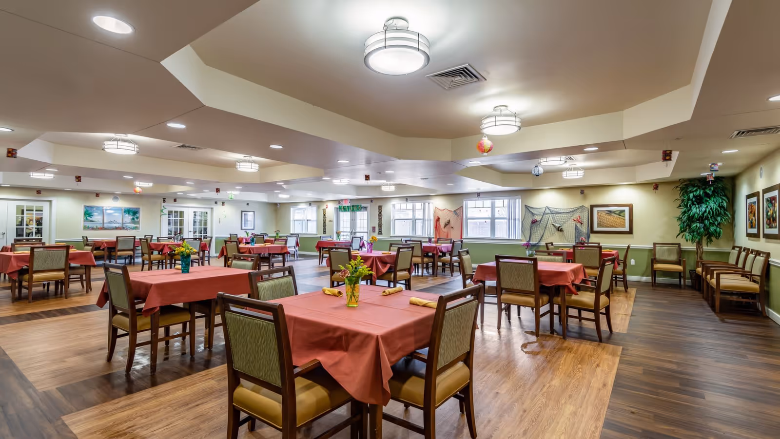 A spacious dining room with multiple tables covered in red tablecloths, each set with yellow napkins and small flower arrangements. The room has wooden flooring, green and beige walls, ceiling lights, and windows letting in natural light. There are framed pictures and decorative nets on the walls, and additional chairs lined up along the wall.