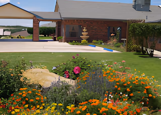 Colorful flower garden and lawn in front of a brick building entrance with a covered driveway and a fountain.