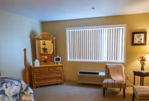 A cozy bedroom with a wooden dresser and mirror, a striped chair with a beige throw blanket, a window with vertical blinds, a floral bedspread partially visible, a small side table with a vase of flowers, and a framed picture on the wall.