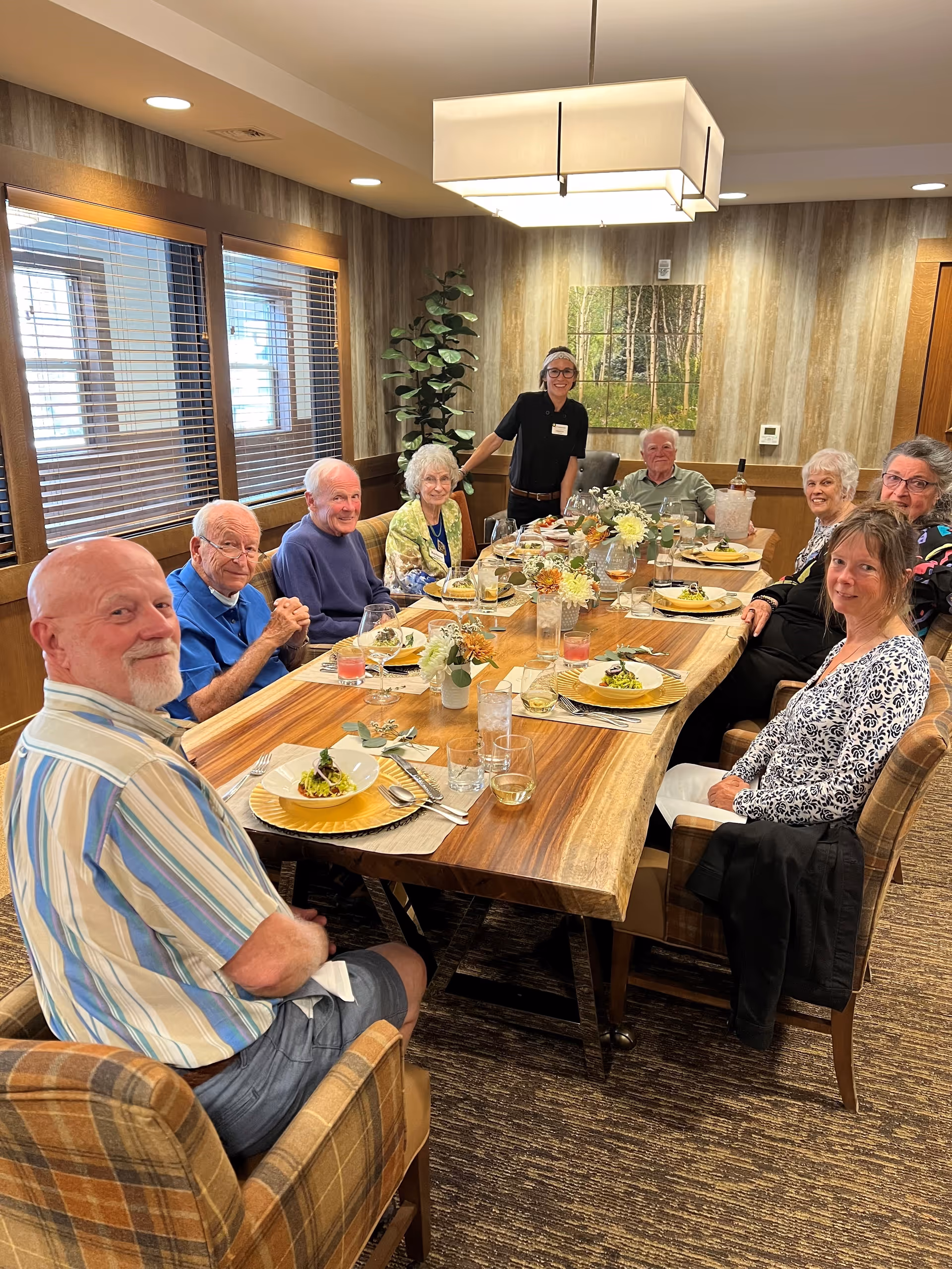 A group of elderly people sitting around a wooden dining table in a warmly lit room, with plates of food and drinks in front of them. A staff member stands smiling behind the table. The room has wood-paneled walls, a large window with blinds, a potted plant, and a nature-themed painting on the wall.
