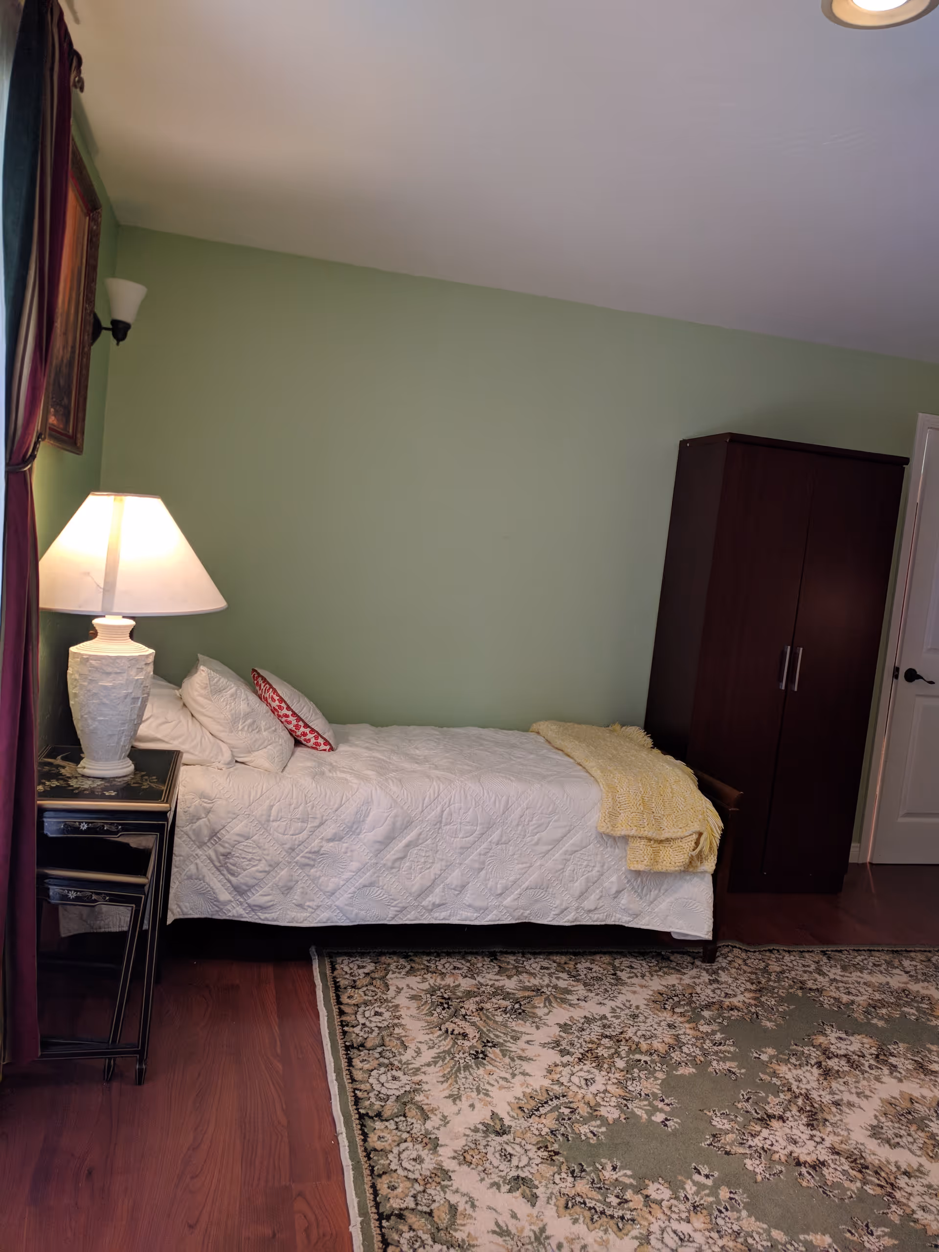 A bedroom with a single bed covered in a white quilt and several pillows, including one with a red pattern. A yellow knitted blanket is folded at the foot of the bed. Next to the bed is a black nightstand with a white lamp on it. The walls are painted light green, and there is a dark wooden wardrobe against the wall. A floral patterned rug covers part of the wooden floor.