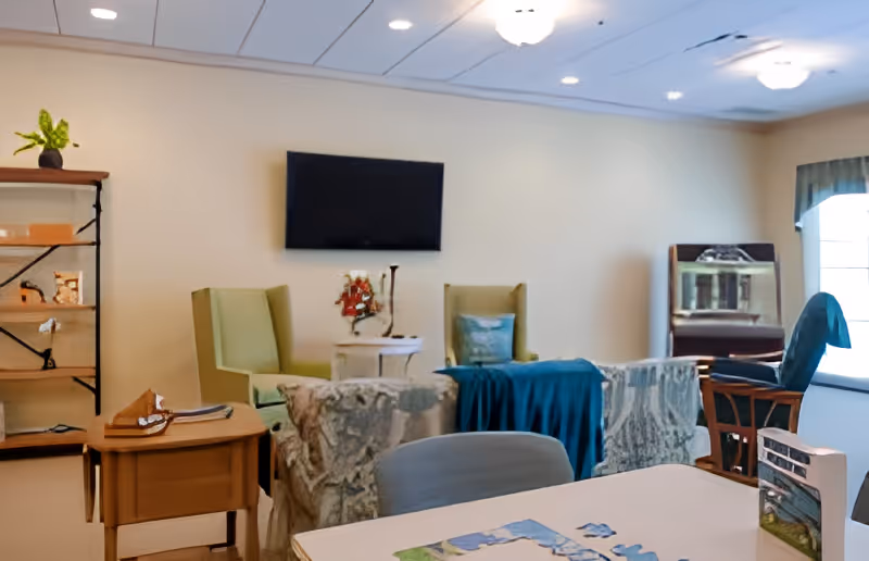A senior living common room with armchairs, a wall-mounted TV, tables, a bookshelf, and a window.