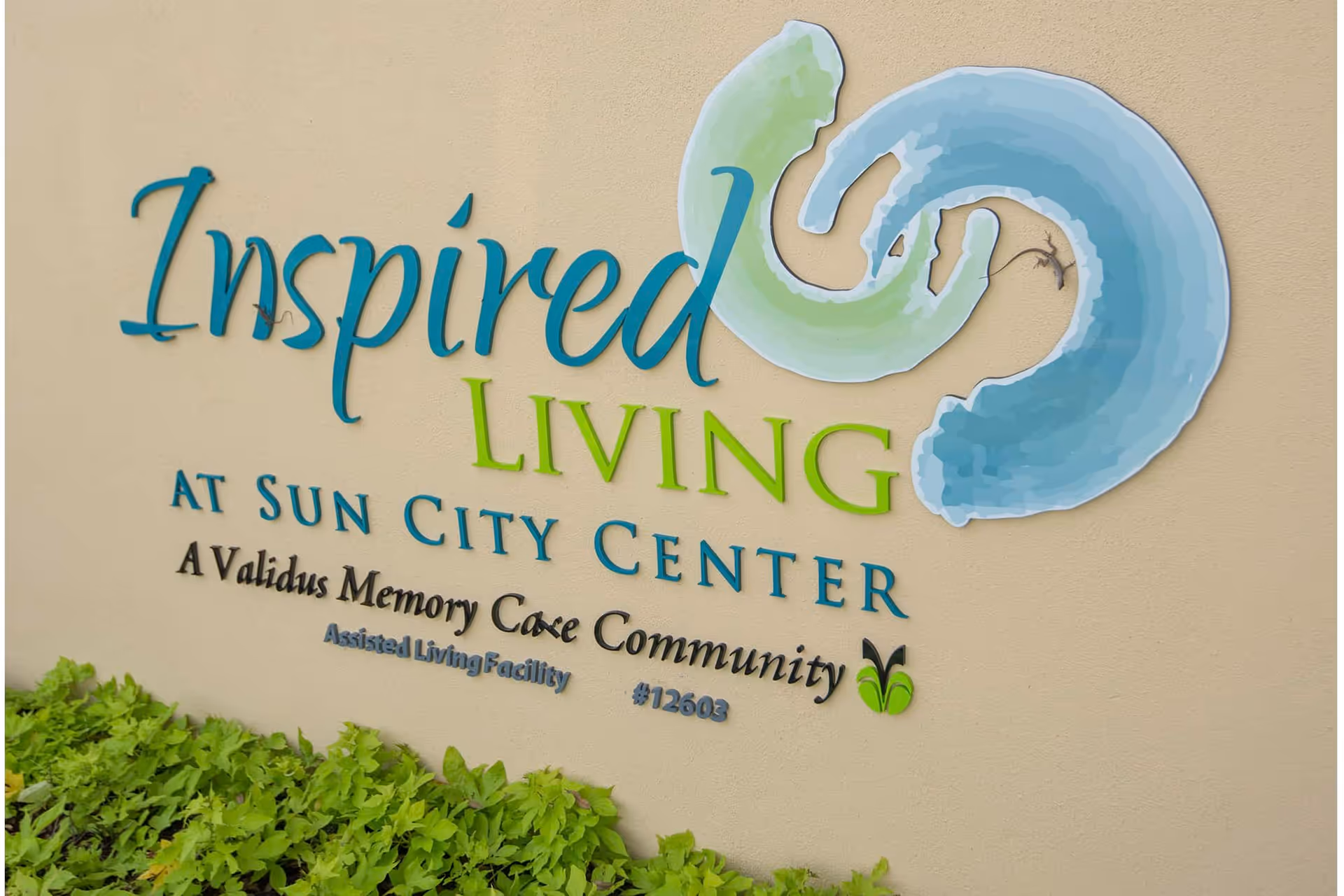 Wall-mounted sign reading "Inspired Living at Sun City Center" with a blue-green logo above low green plants.