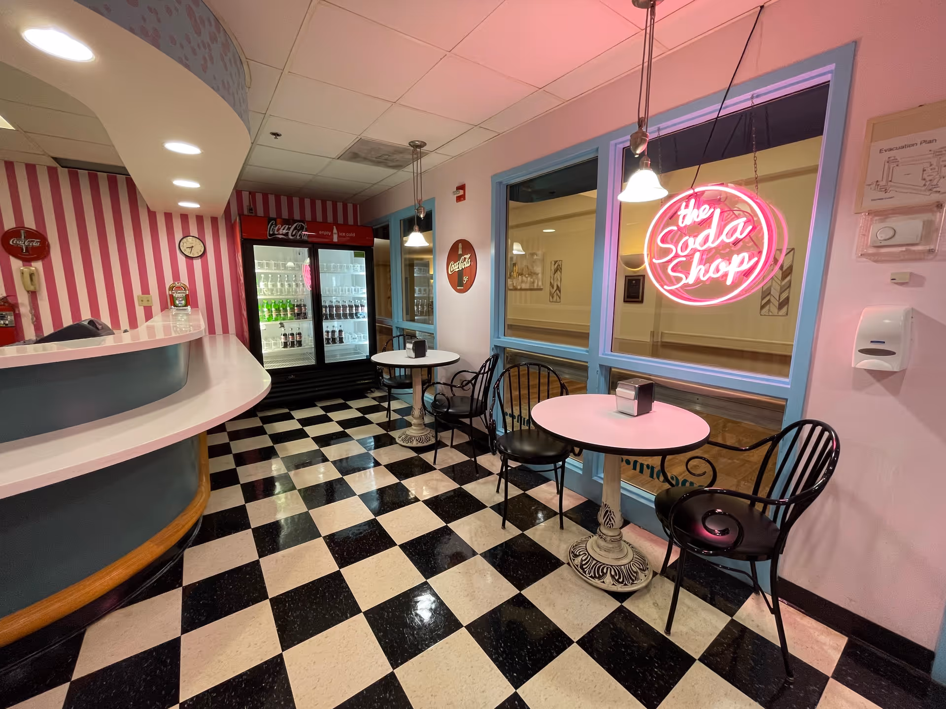 Retro soda shop-style seating area with a black-and-white checkered floor, pink-striped counter, refrigerated drink case, round tables and a neon 'the Soda Shop' sign.