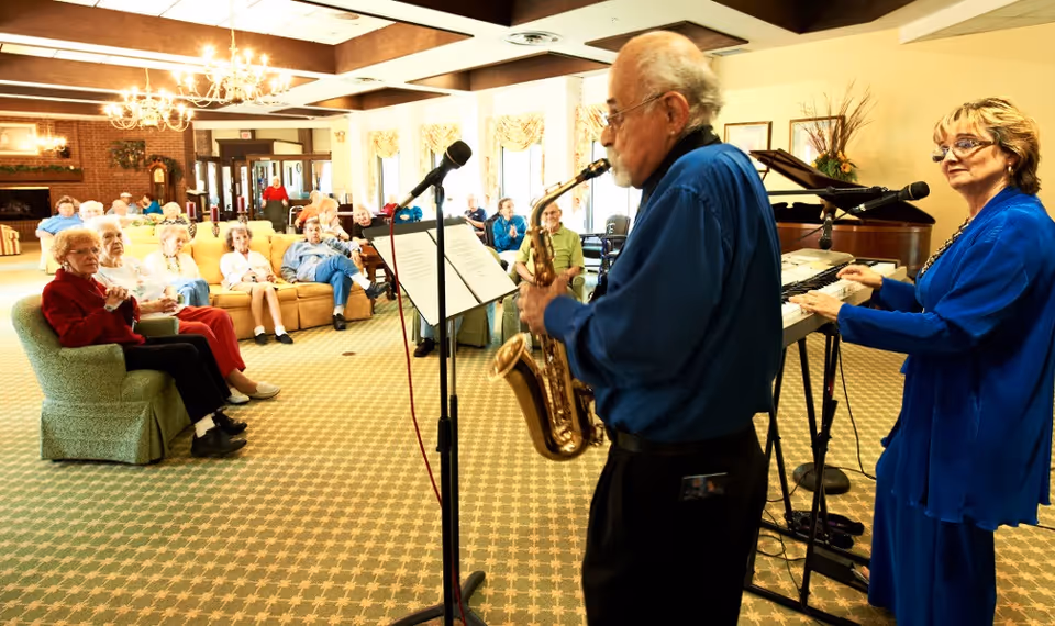 Musicians playing saxophone and keyboard performing for a seated group of elderly residents in a bright communal living room.