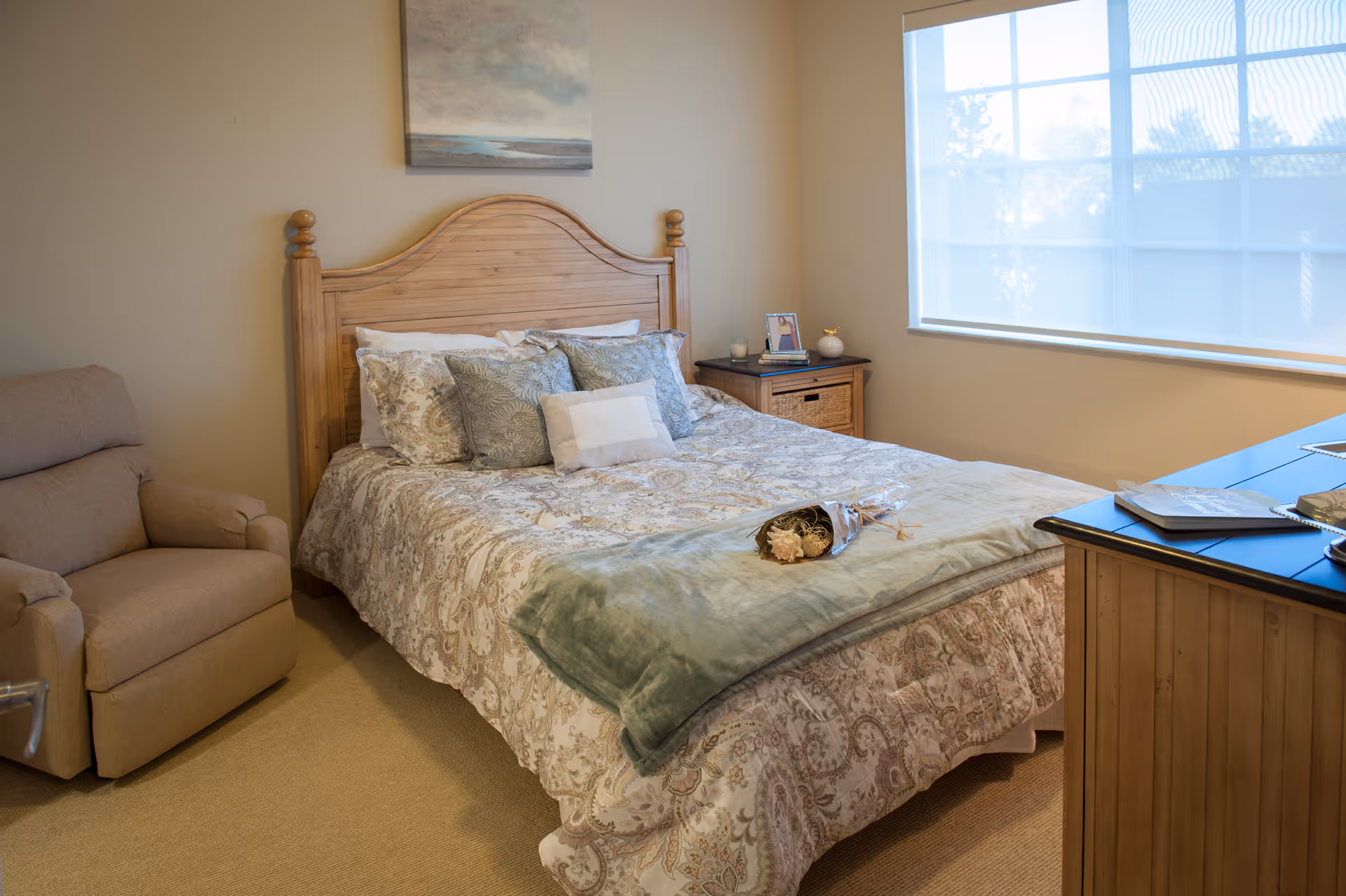 A cozy bedroom with a wooden headboard bed covered in patterned bedding and multiple pillows. A beige recliner chair is positioned to the left of the bed. There is a wooden nightstand with a small framed photo, a candle, and a decorative item. A large window with a white shade allows natural light into the room. A wooden dresser with books and a decorative tray is on the right side of the image.