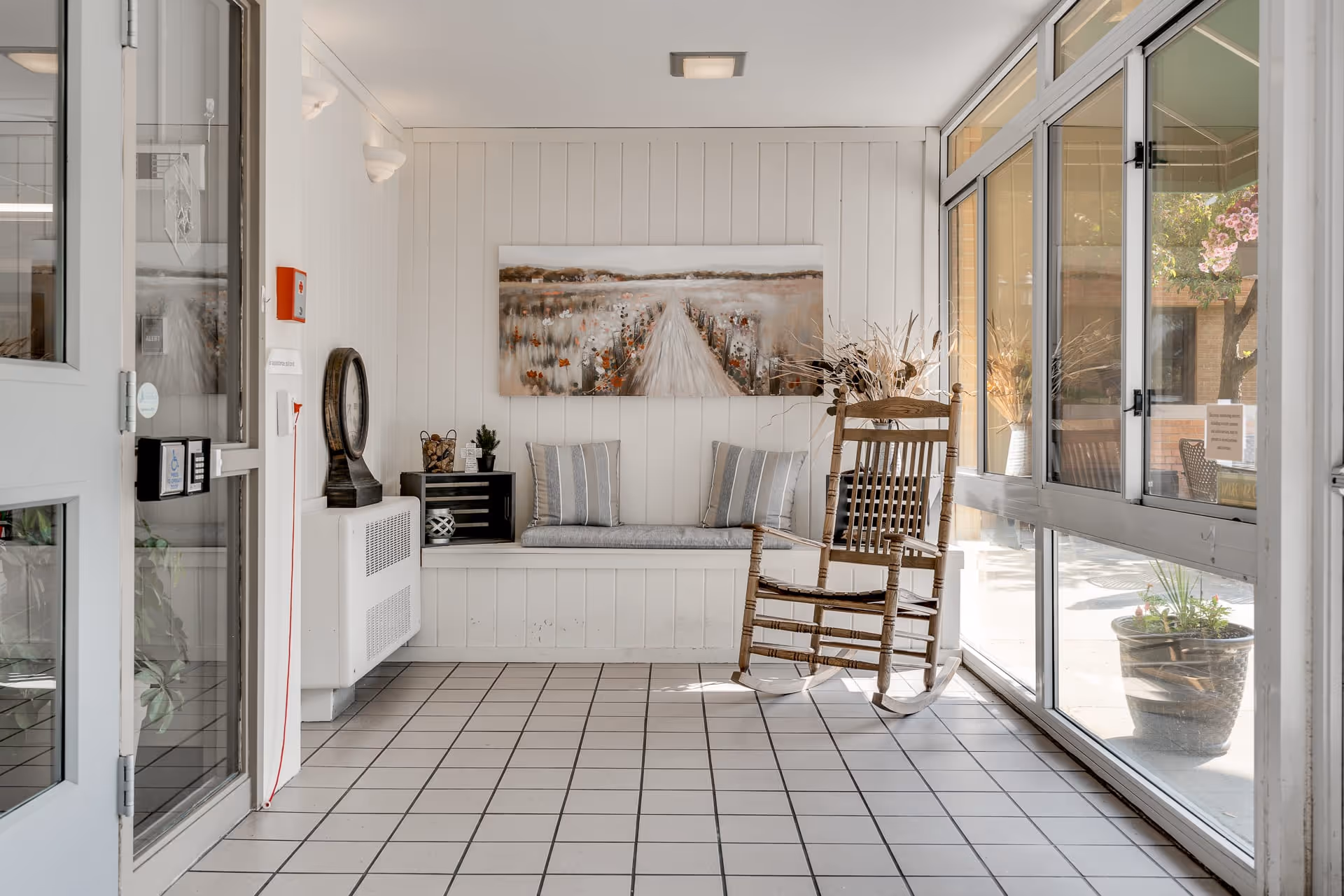 A bright indoor seating area with white paneled walls and tiled floor. There is a wooden rocking chair on the right side near large windows, and a built-in bench with two striped cushions beneath a landscape painting of a path through a field. Decorative items including a clock, small plants, and dried flowers are arranged on and around the bench.