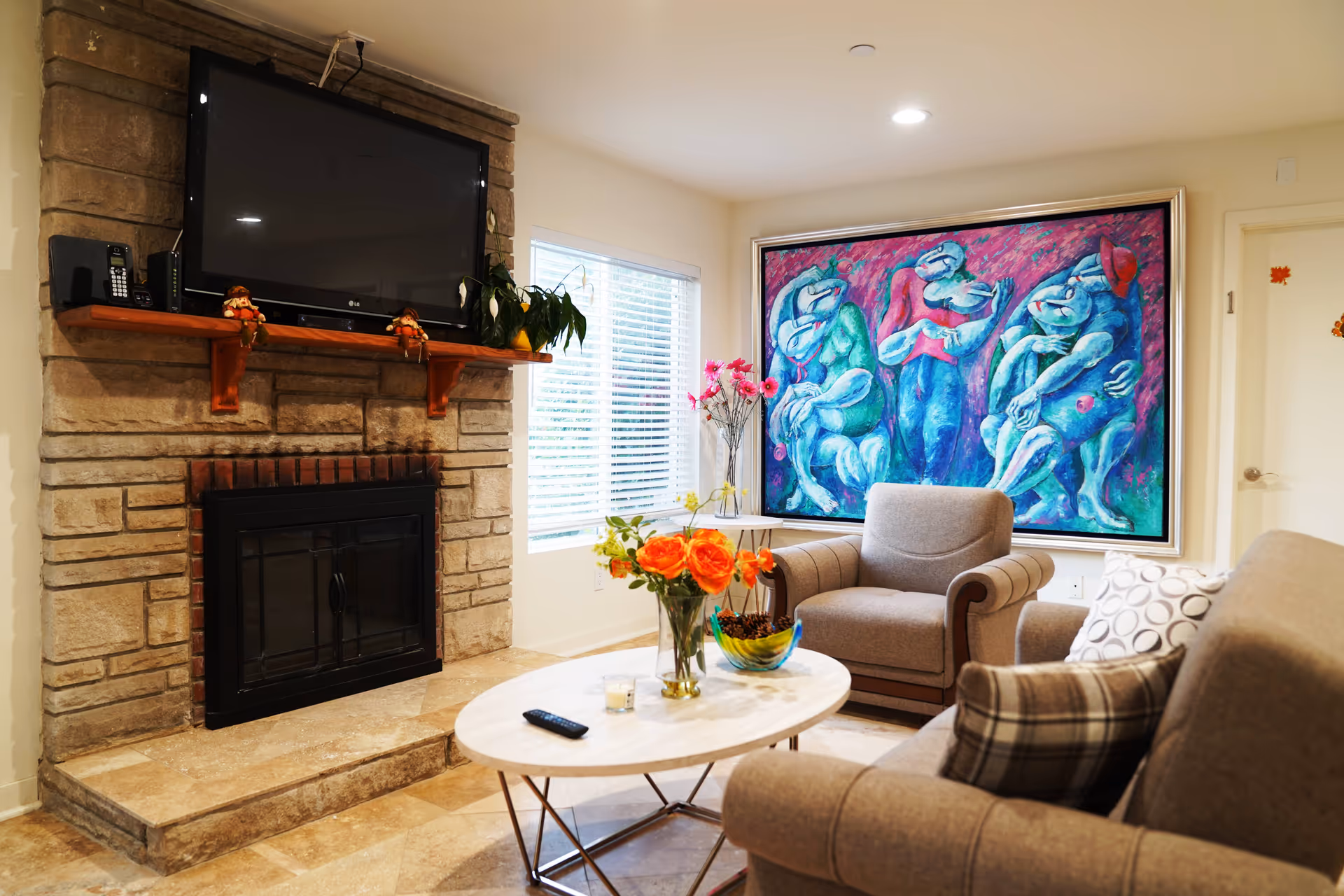 A cozy living room with a stone fireplace and a mounted flat-screen TV above it. There is a wooden mantel with decorative items and a potted plant. The room has a large colorful painting on the wall, two comfortable armchairs, a sofa with patterned pillows, and a white oval coffee table with a vase of orange flowers, a candle, a remote control, and a decorative bowl. A window with blinds allows natural light into the room.