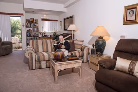 An elderly man sits on a striped sofa reading a book in a carpeted living room with lamps, armchairs, and bookshelves.