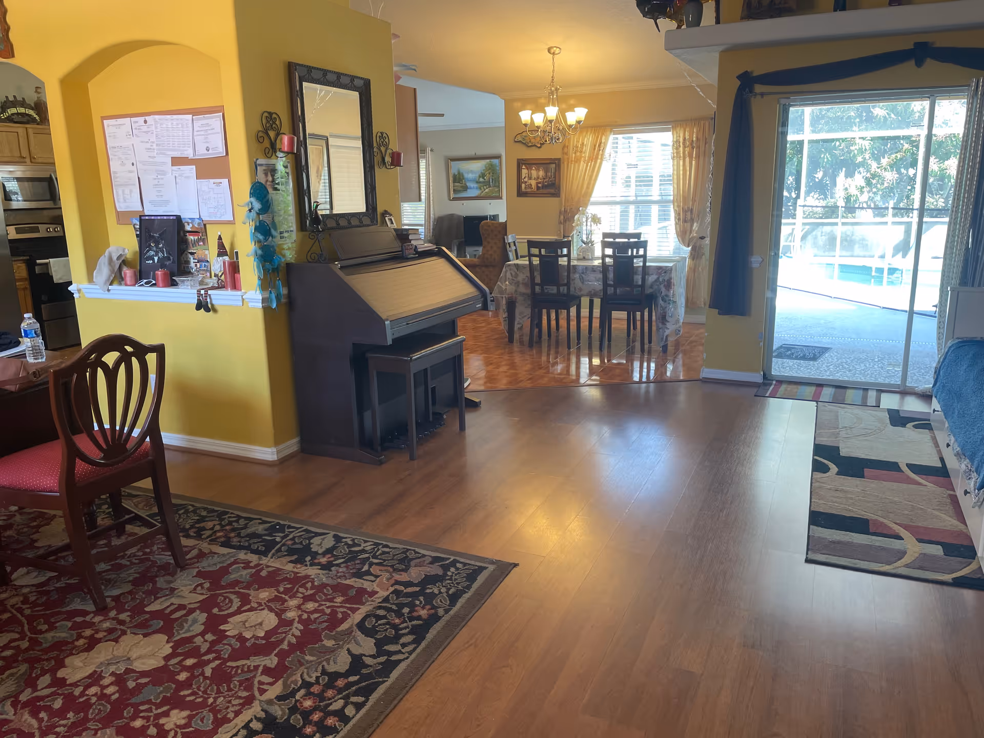 Bright yellow interior with piano, bulletin board, dining table, and sliding glass door.