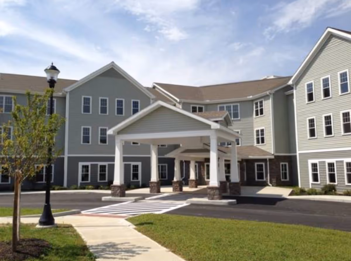 Front entrance of a three-story assisted living building with a covered porte-cochere, driveway, and lawn.