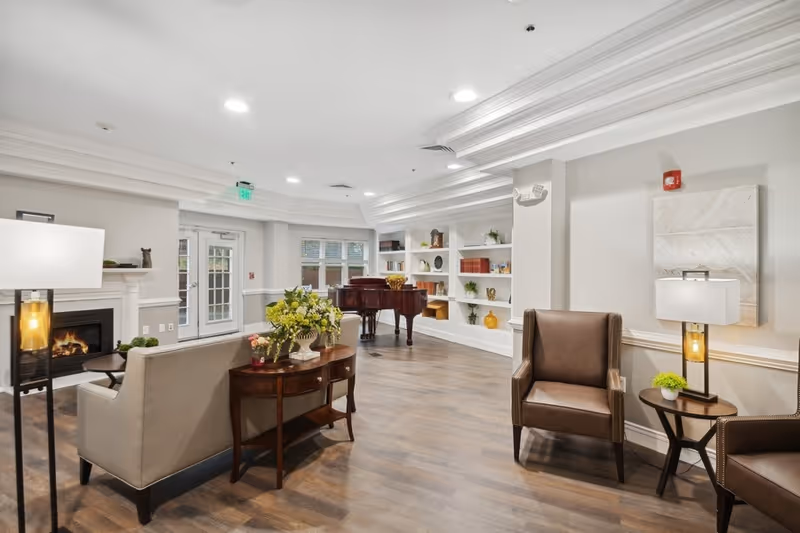A bright and spacious living room area with wooden flooring, a beige sofa, two brown leather armchairs, a wooden side table with a lamp, a fireplace, built-in white shelves with decorative items, and a grand piano near the windows.