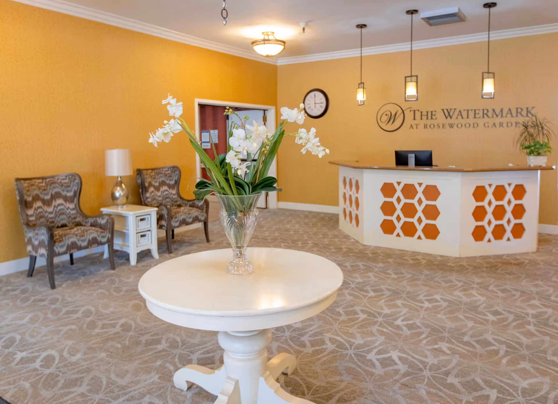 Reception area of The Watermark at Rosewood Gardens featuring a white round table with a glass vase of white flowers in the foreground, two patterned armchairs with a small white side table and lamp against a yellow wall, and a reception desk with pendant lights and a clock on the wall behind it.