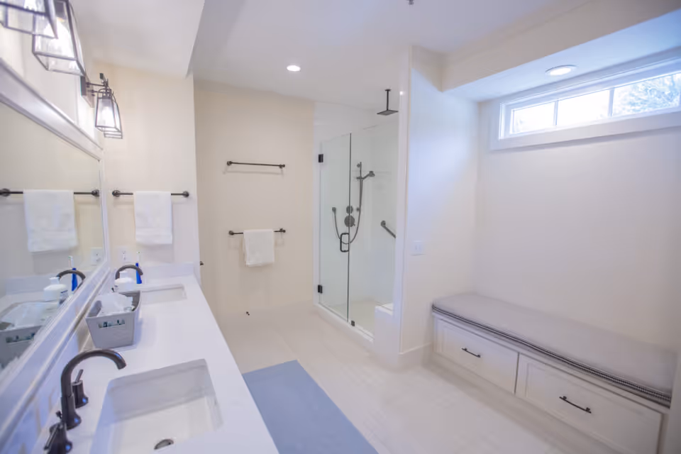 Bright and modern bathroom featuring a double sink vanity with dark faucets, a large mirror, white towels hanging on black towel racks, a glass-enclosed shower with a rain showerhead, and a cushioned bench with storage drawers beneath a high window.