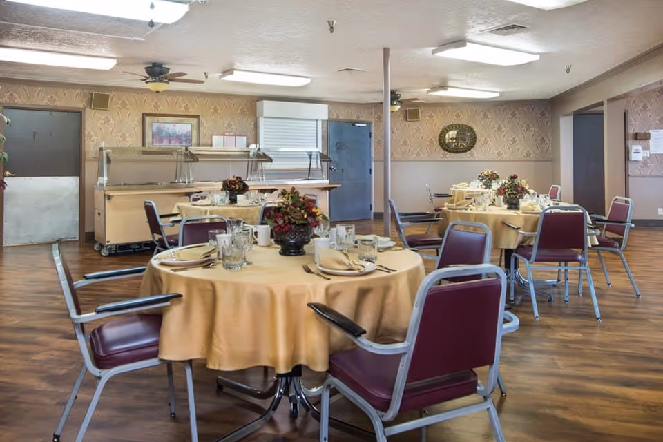 Dining room with round tables covered in beige tablecloths, each set with plates, glasses, napkins, and floral centerpieces. The room has wooden flooring, patterned wallpaper, ceiling fans, and a serving area with a metal counter and doors in the background.