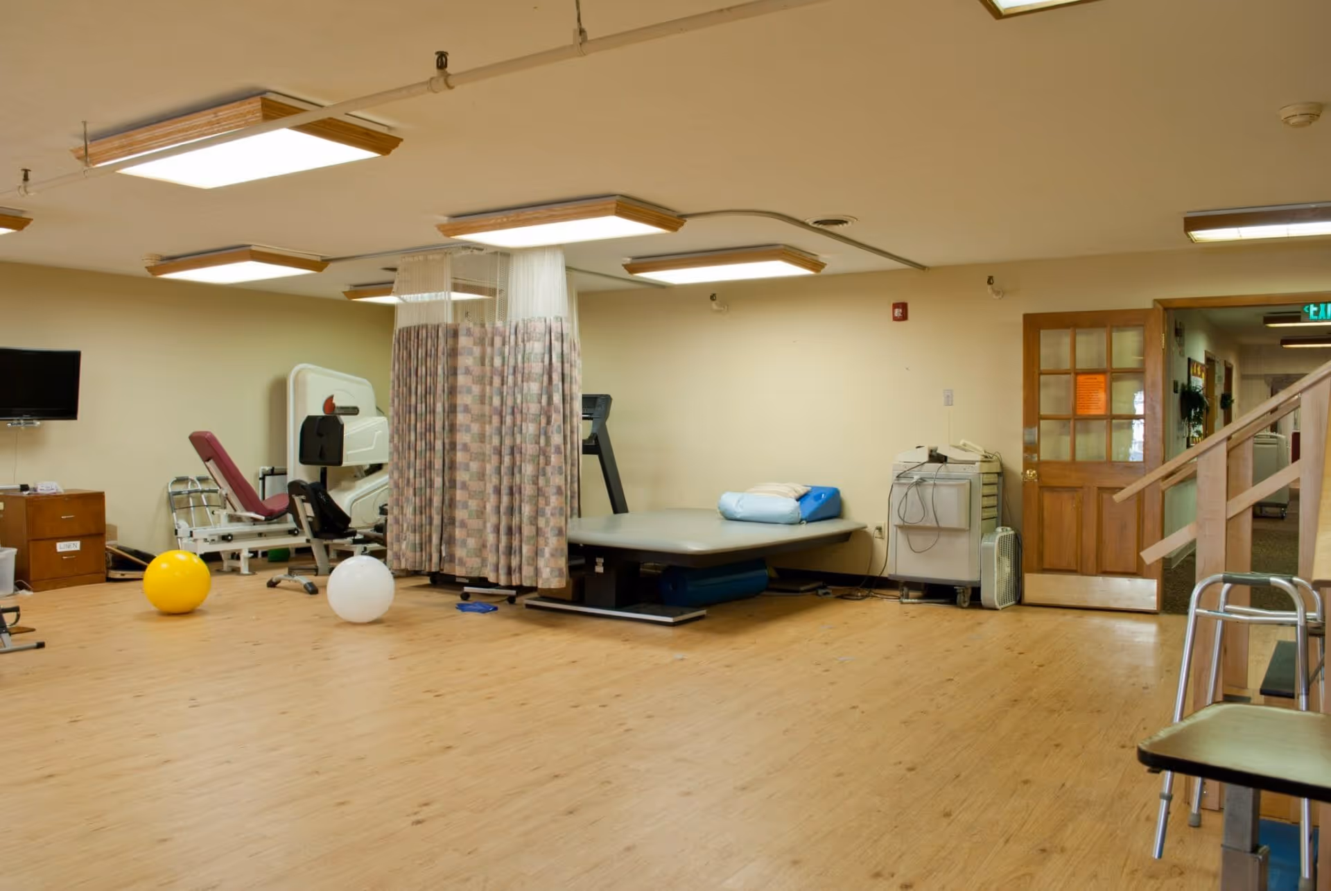 Spacious therapy/exercise room with a treatment table, exercise equipment, therapy balls, privacy curtains, and wood flooring.