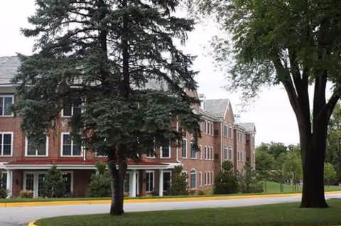 Brick senior living building exterior with trees, curved driveway, and manicured lawn.
