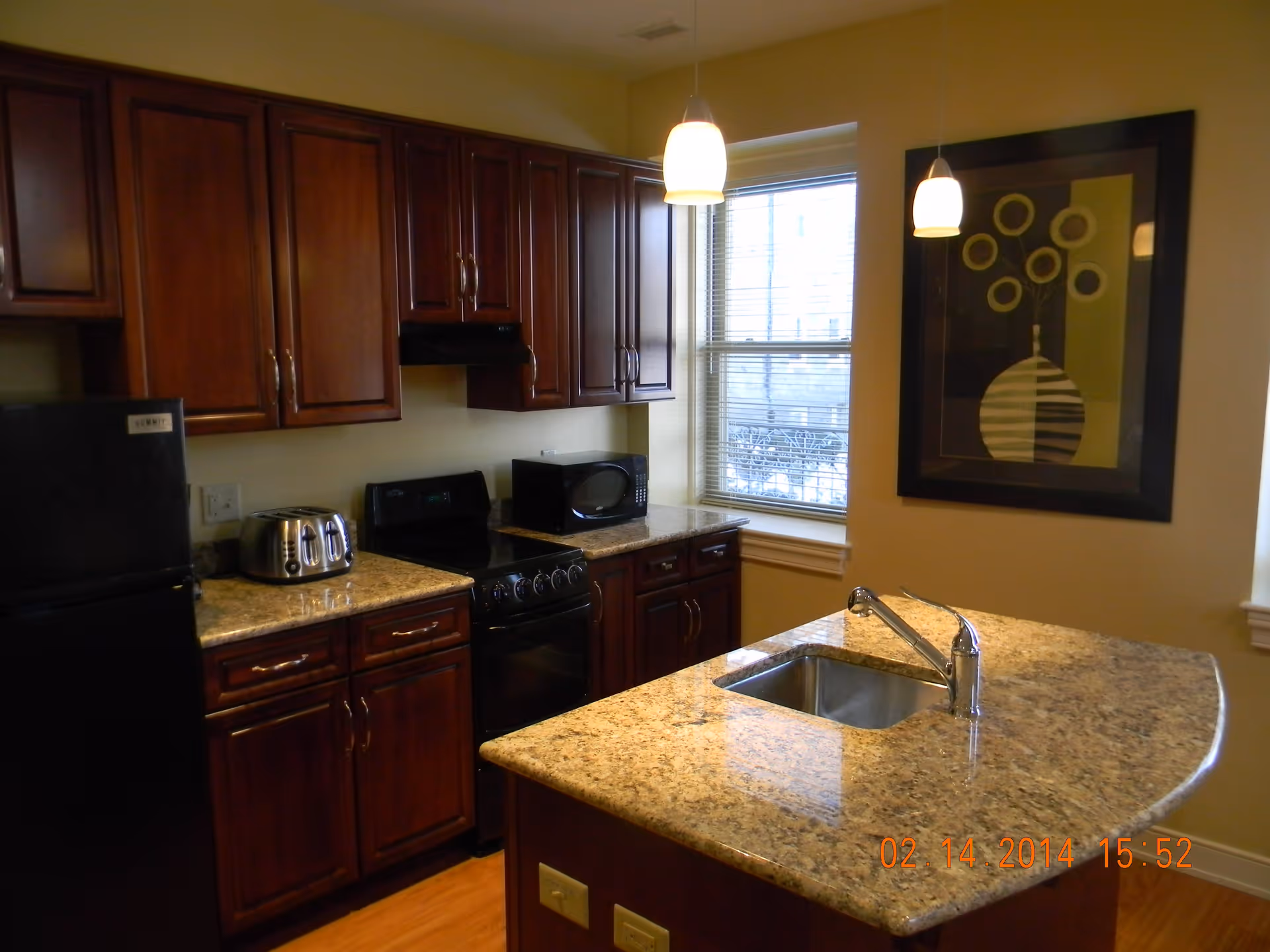 A kitchen with dark wooden cabinets, a black refrigerator, a black stove, a toaster, a microwave, and a granite countertop island with a sink. Two pendant lights hang from the ceiling, and there is a window with blinds and a framed abstract artwork on the wall.