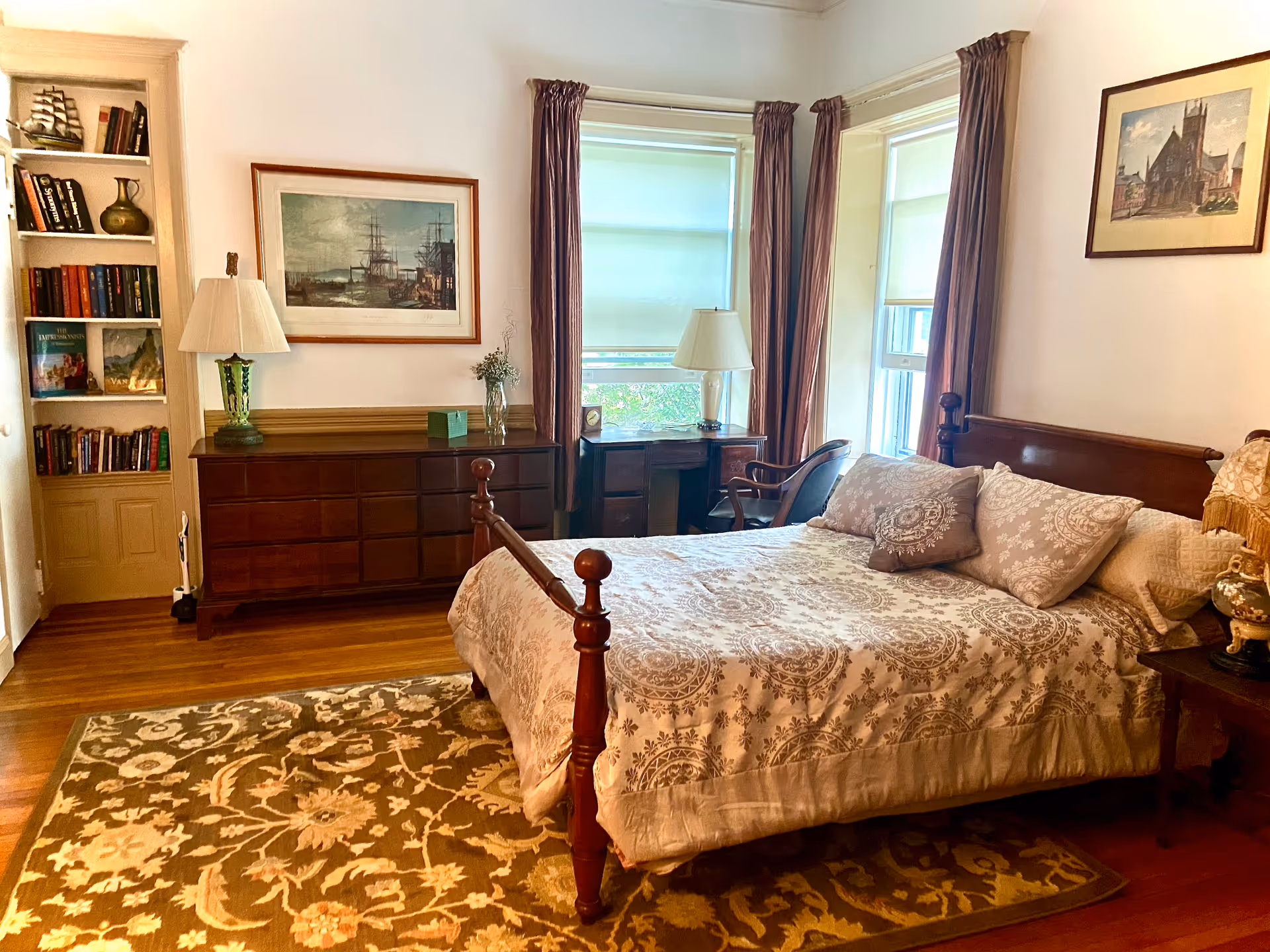 A cozy bedroom with a wooden bed featuring patterned bedding and multiple pillows. There is a wooden dresser with a lamp and decorative items, a small desk with a chair near two windows with curtains and blinds, and a built-in bookshelf filled with books. The room has wooden flooring and a patterned area rug.