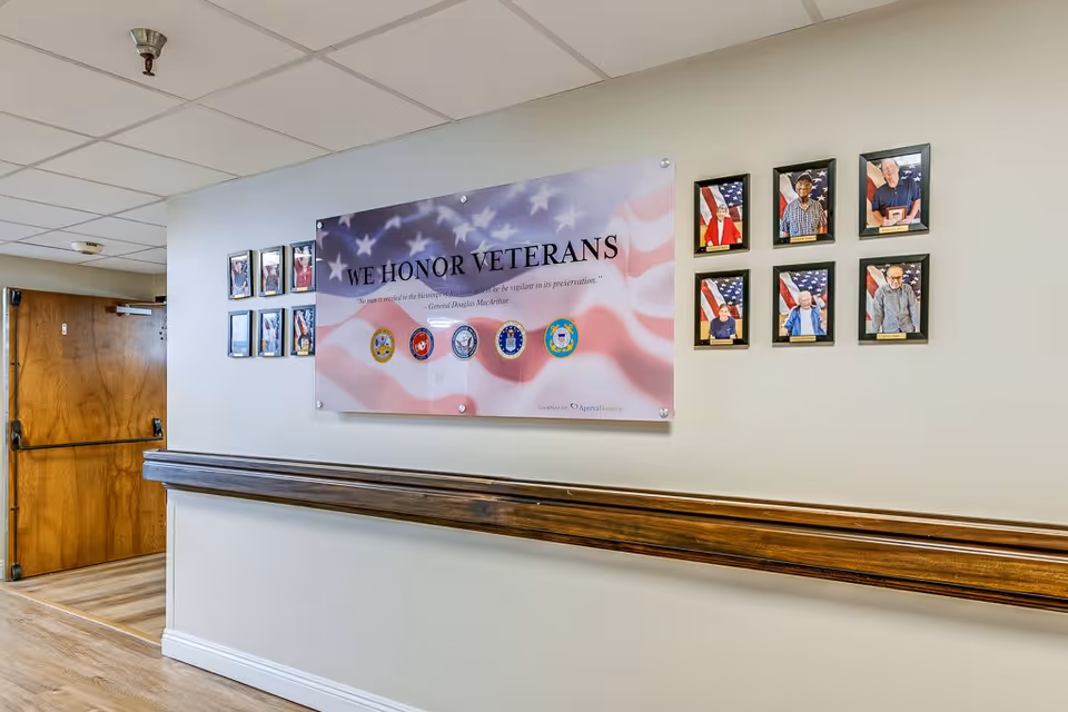 A hallway in a senior living facility with a wooden handrail along the wall. On the wall, there is a large sign that reads 'WE HONOR VETERANS' with military service emblems and a quote by General Douglas MacArthur. Surrounding the sign are framed photographs of veterans with American flags in the background. The hallway has a wooden door at the end and a tiled ceiling.